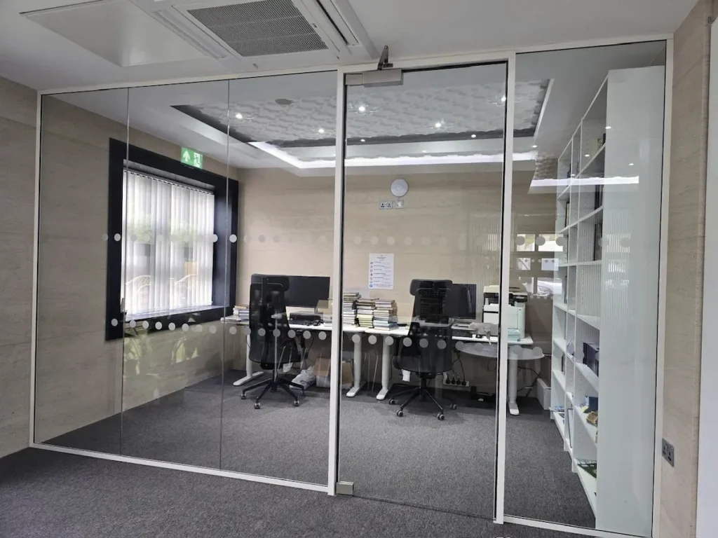 White-framed glass office partition with dotted manifestation, enclosing a desk area with printer and bookshelf visible