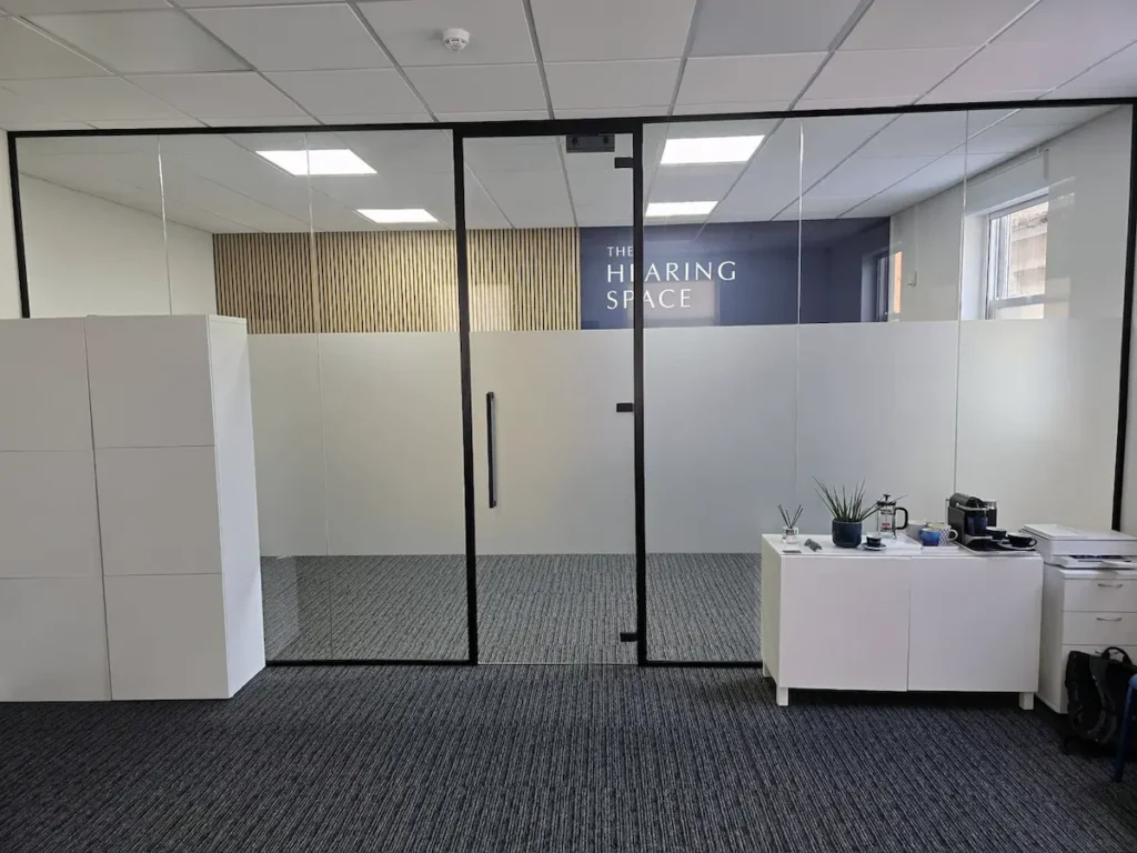 Black-framed glass office partition with a hinged glass door, opening onto a reception area with patterned carpet