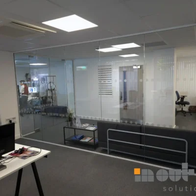 Frameless glass partitions viewed from a desk, dotted manifestation strip and treatment equipment visible through the glass