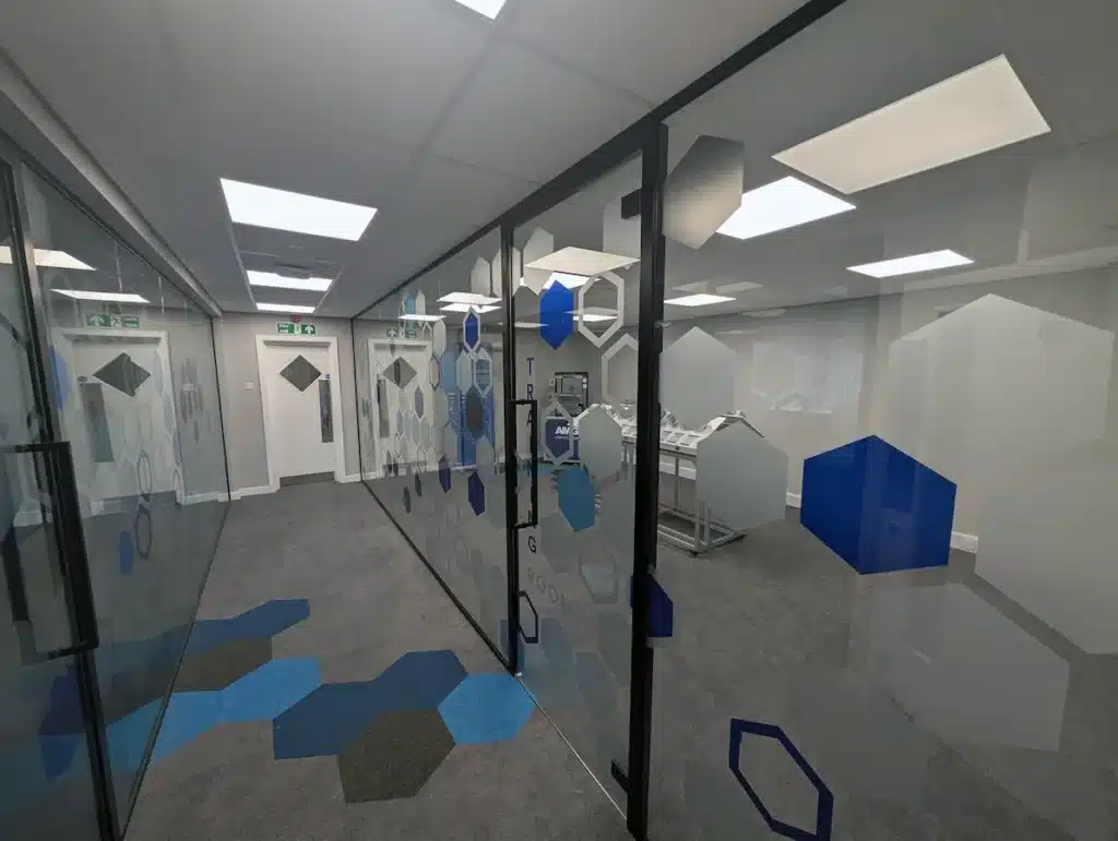 Corridor view of glass partitions with hexagon branding and door handle, exit signage and hexagon-patterned carpet visible