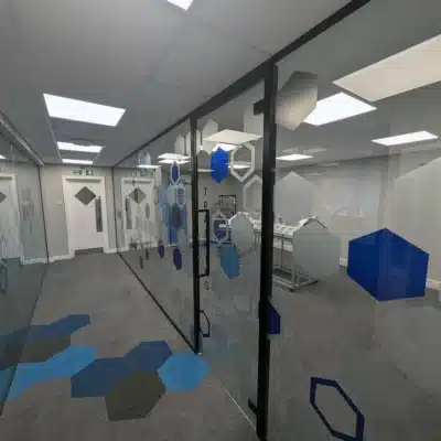 Corridor view of glass partitions with hexagon branding and door handle, exit signage and hexagon-patterned carpet visible