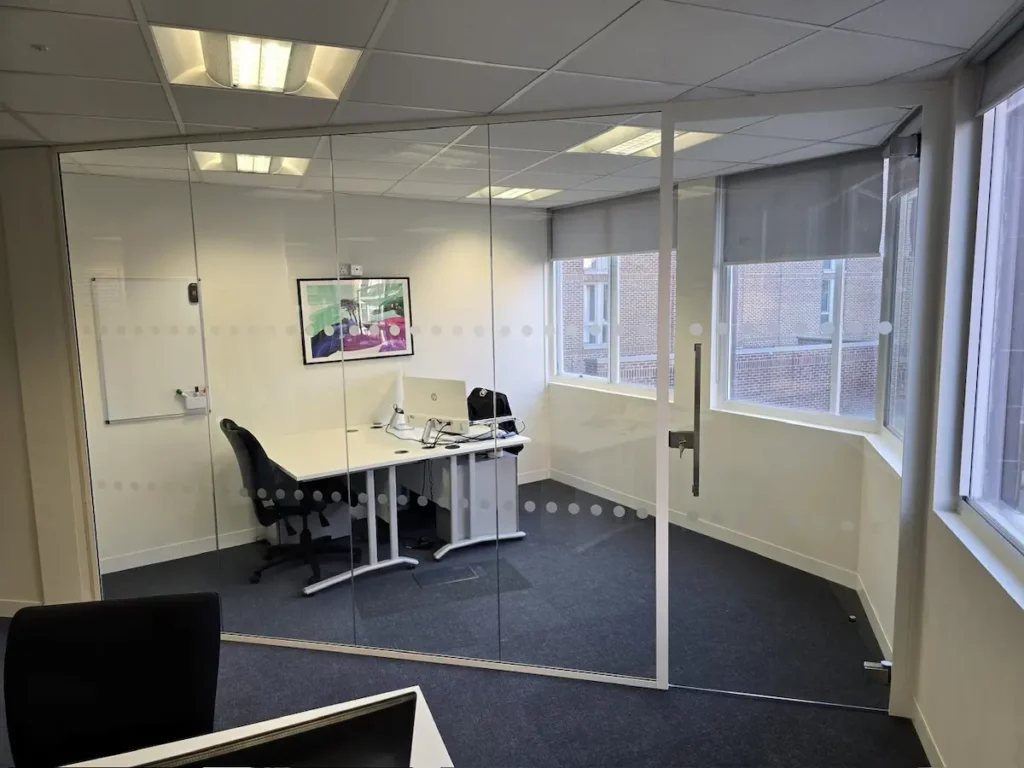 Glass partition viewed from inside a private office, showing a desk, window, and wall-mounted artwork
