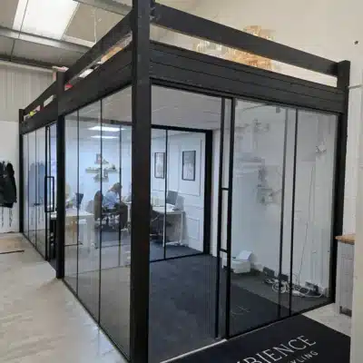 Corner view of a black-framed glass office pod with sliding door, desks visible inside, exposed beams above