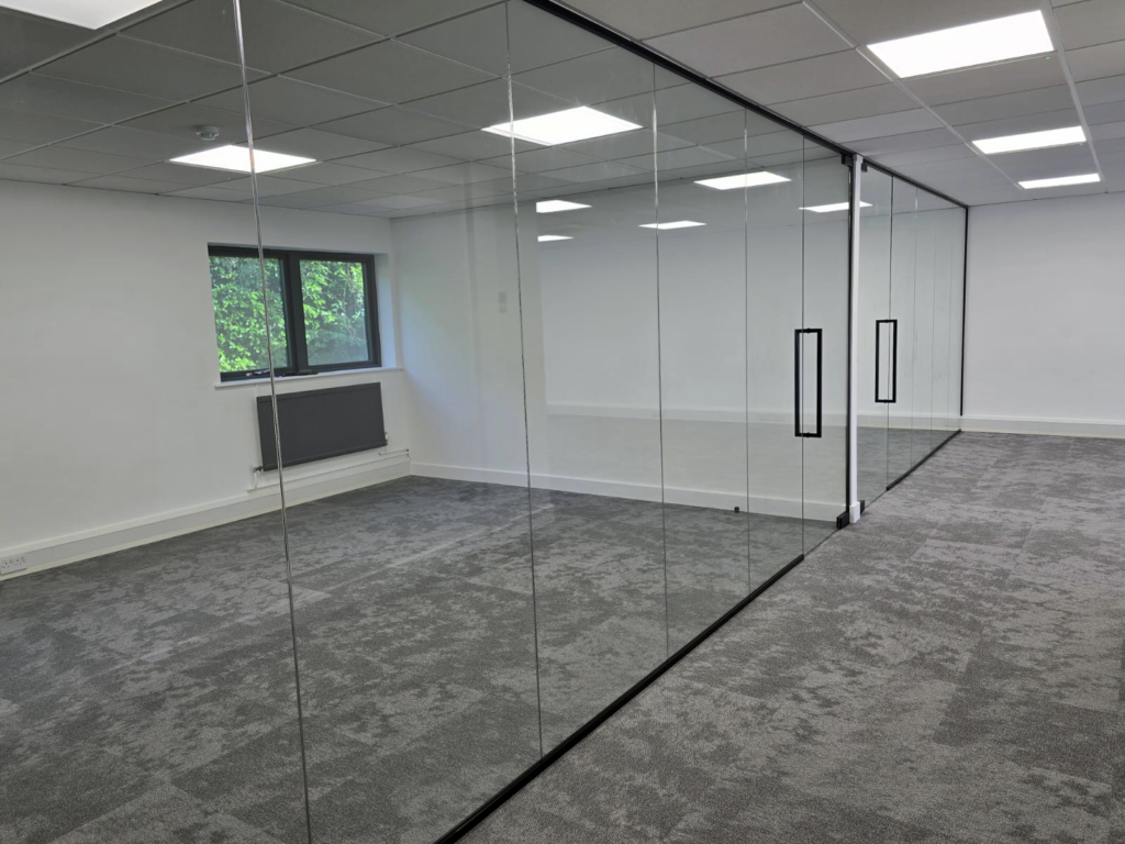 Frameless glass partition with black vertical door handles, corner view of an office with carpet tiles