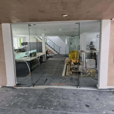 Wider view of a frameless glass partition and sliding door, desks and staircase visible through the glass, construction materials on the floor