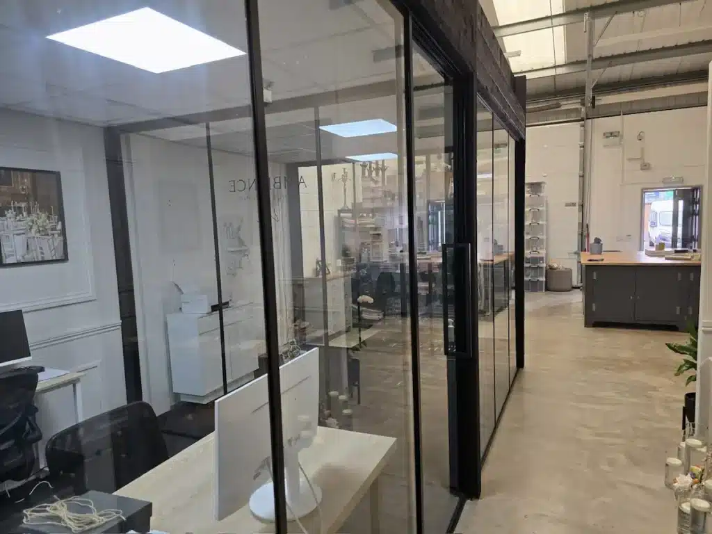 Row of black-framed glass partitions along a corridor in an industrial office, desks and monitors visible through the glass