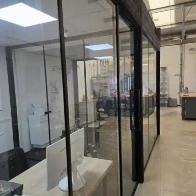 Row of black-framed glass partitions along a corridor in an industrial office, desks and monitors visible through the glass