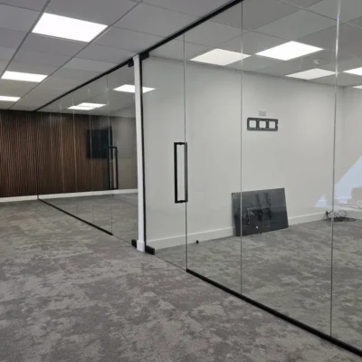 Frameless Glass Partitions. Corner view of glass partitions meeting at a white support column, wood-slat wall and mounted screen visible beyond