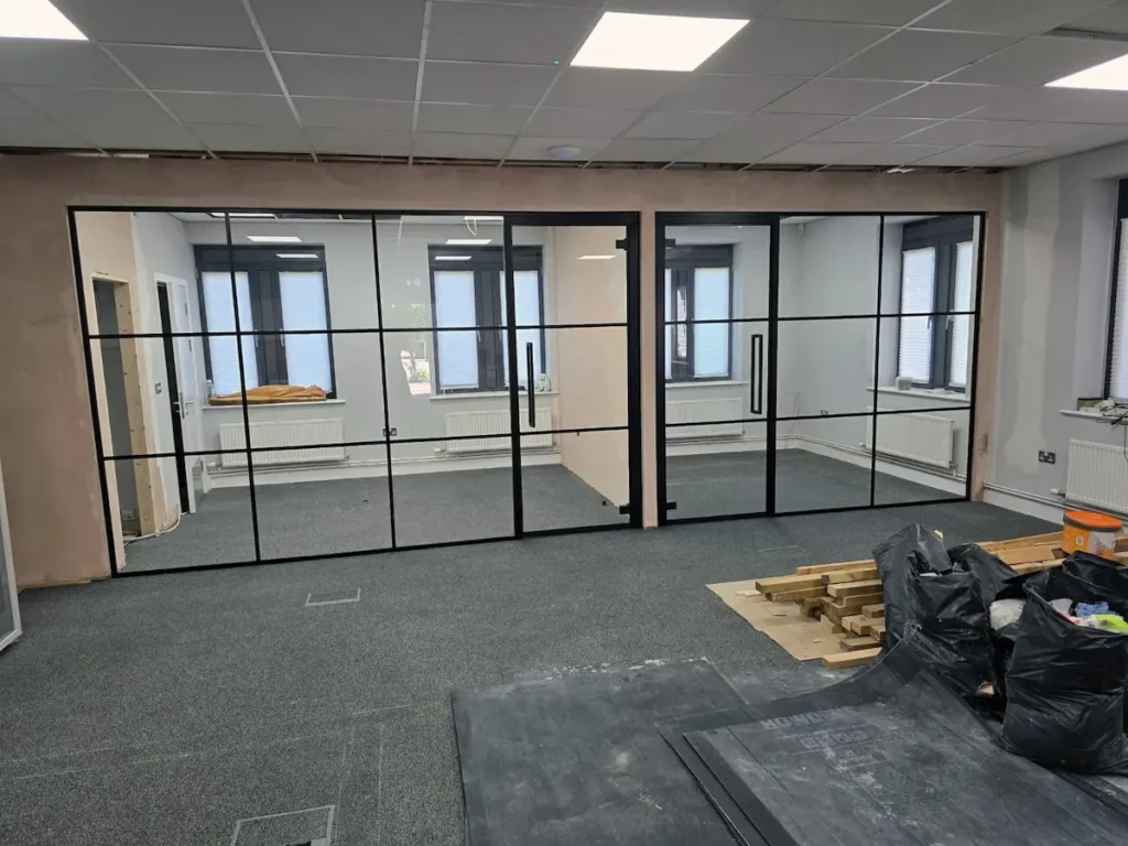 Wide view of steel-framed glass partitions dividing two adjoining offices in Bath, windows and construction materials visible