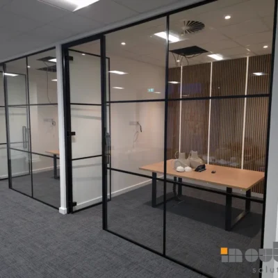 Corner view of a black aluminium grid-framed glass partition enclosing a desk area, wood-panel wall visible through the glass