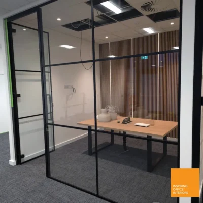 Black aluminium grid-framed glass partition with open hinged door, desk and wood-panel wall visible inside
