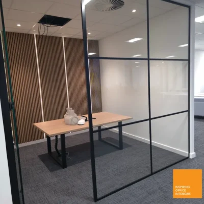 Black aluminium grid-framed glass partition with hinged door open, desk and wood-slat wall visible inside a private office pod