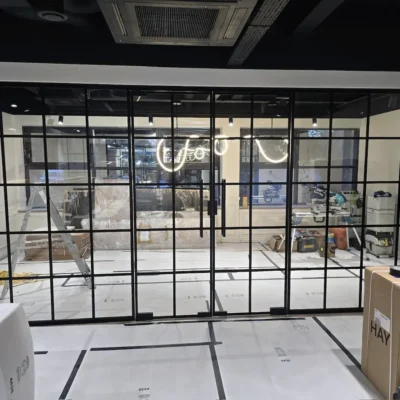 Black framed glass partition and door viewed from outside, with neon signage visible through the glass