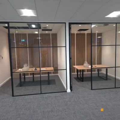 Two adjoining glazed office pods with aluminium grid framing, desks and wood-slat walls visible, hinged doors on each pod