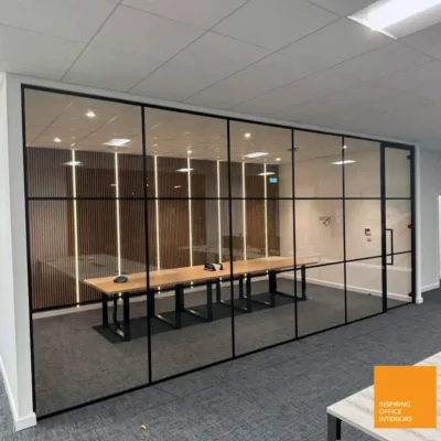 Black-grid glass partition dividing an open-plan office with wood-slat feature wall, desks visible through the glazing