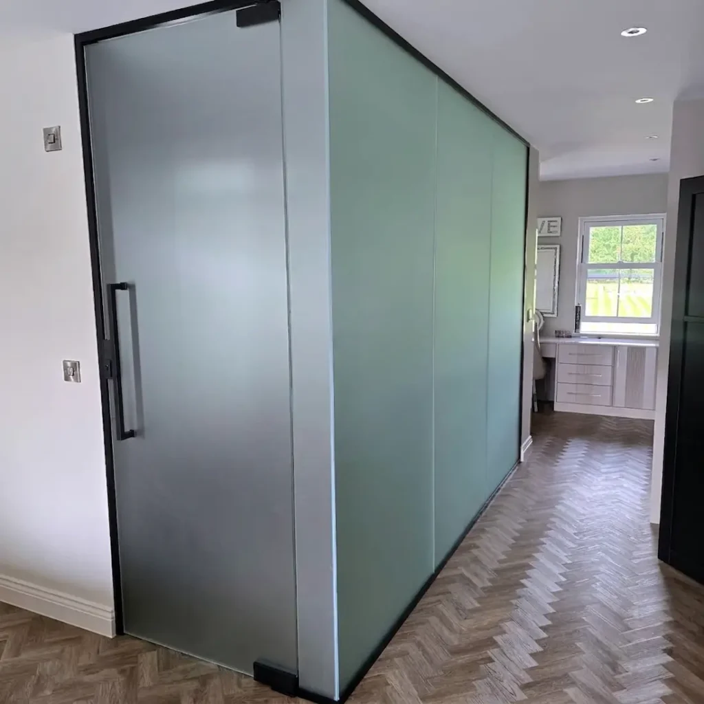 Frosted glass partition with black-framed aluminium hinged door, enclosing an en-suite bathroom off a hallway with herringbone flooring