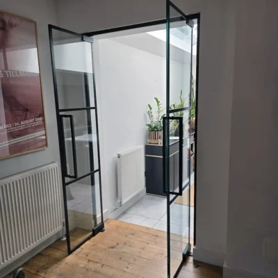 Black-framed glass door open on a hinge, revealing a kitchen with tiled flooring and radiator
