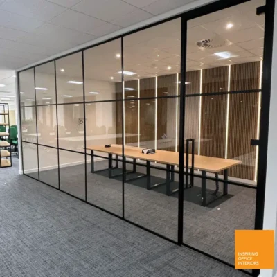 Black aluminium grid-framed glass partition with hinged door, angled view of an office with wood-slat wall and desk seating